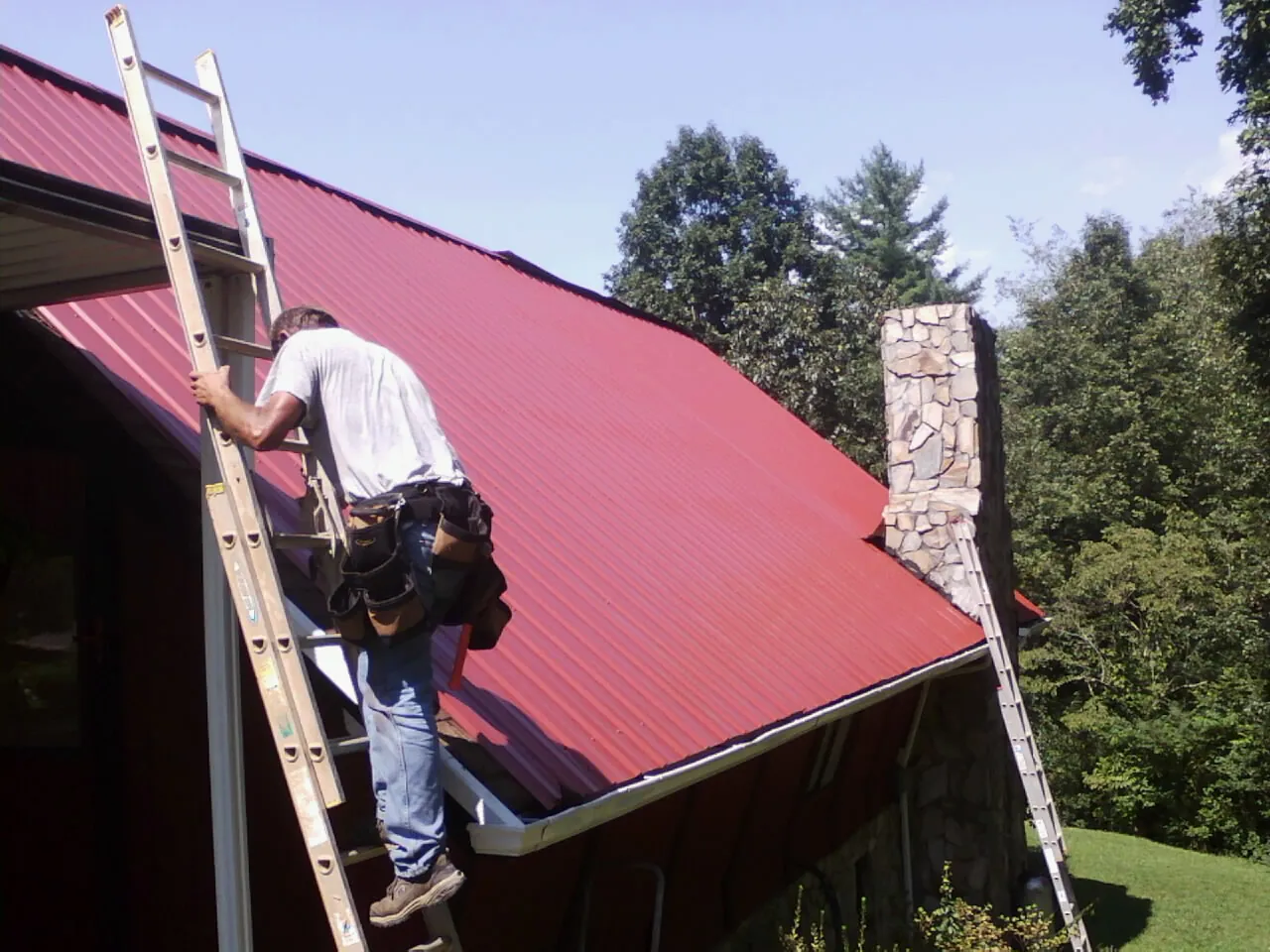Roofer on a ladder beside a red metal roof and stone chimney — Soffit & Fascia Repair by Roofing Brothers in Red Hook